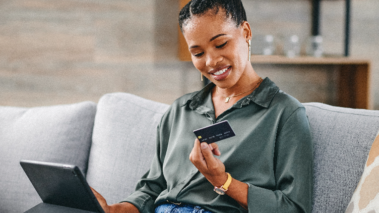 Woman sitting on couch, wearing green blouse and blue jeans holding tablet on her lap and looking at the back of her bank card.